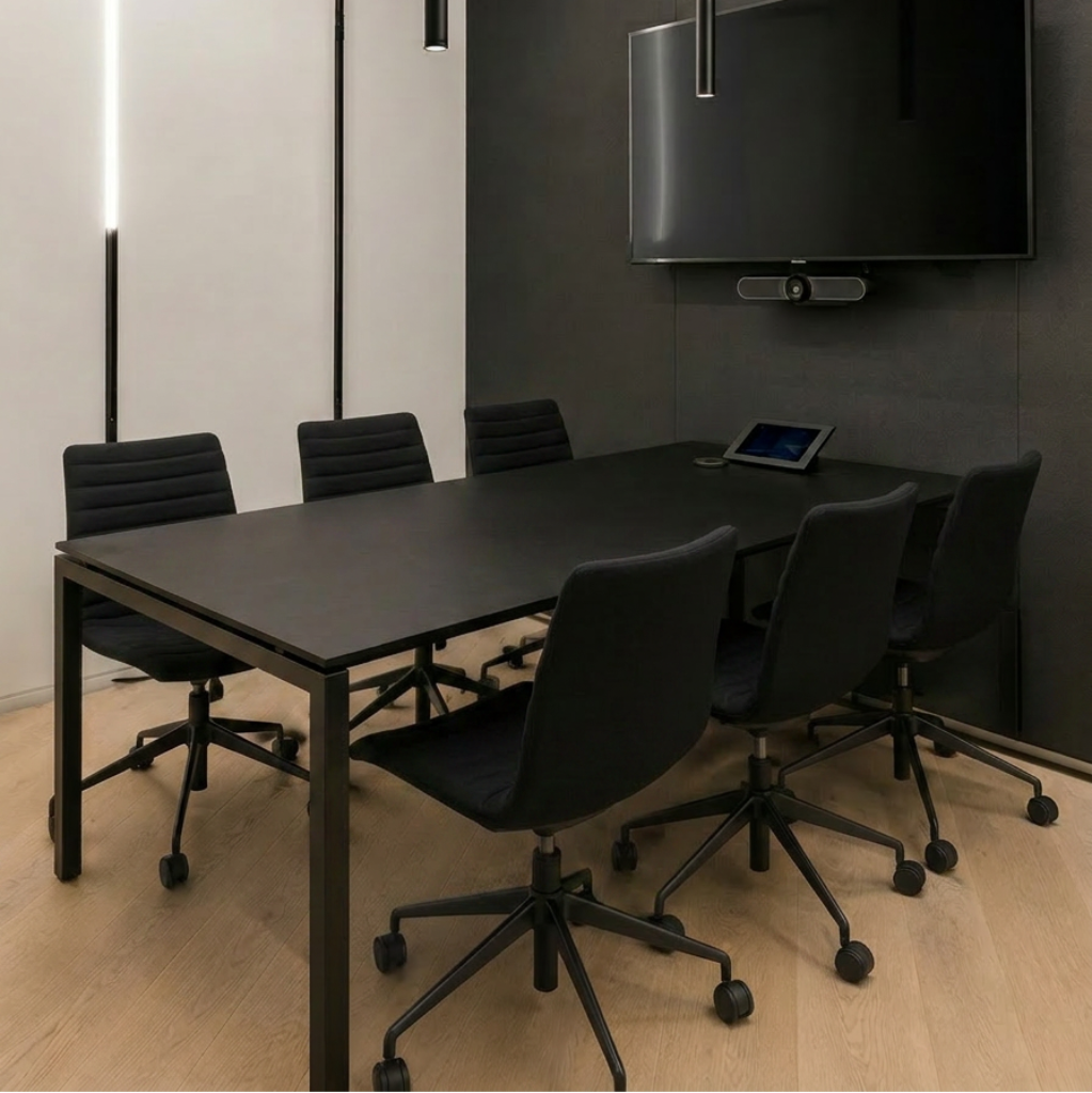 Modern conference room setup with a black rectangular meeting table, six black office chairs, a wall-mounted flat screen TV, and sleek black pendant lights over a light wood floor.