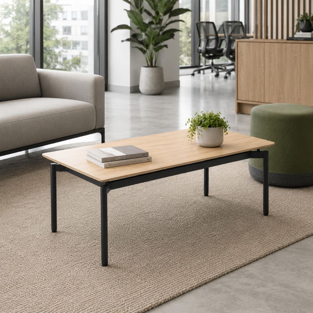 A rectangular coffee table with a light natural wood top and black metal legs, styled in a modern office lounge setting with a sofa, rug, and potted plant.