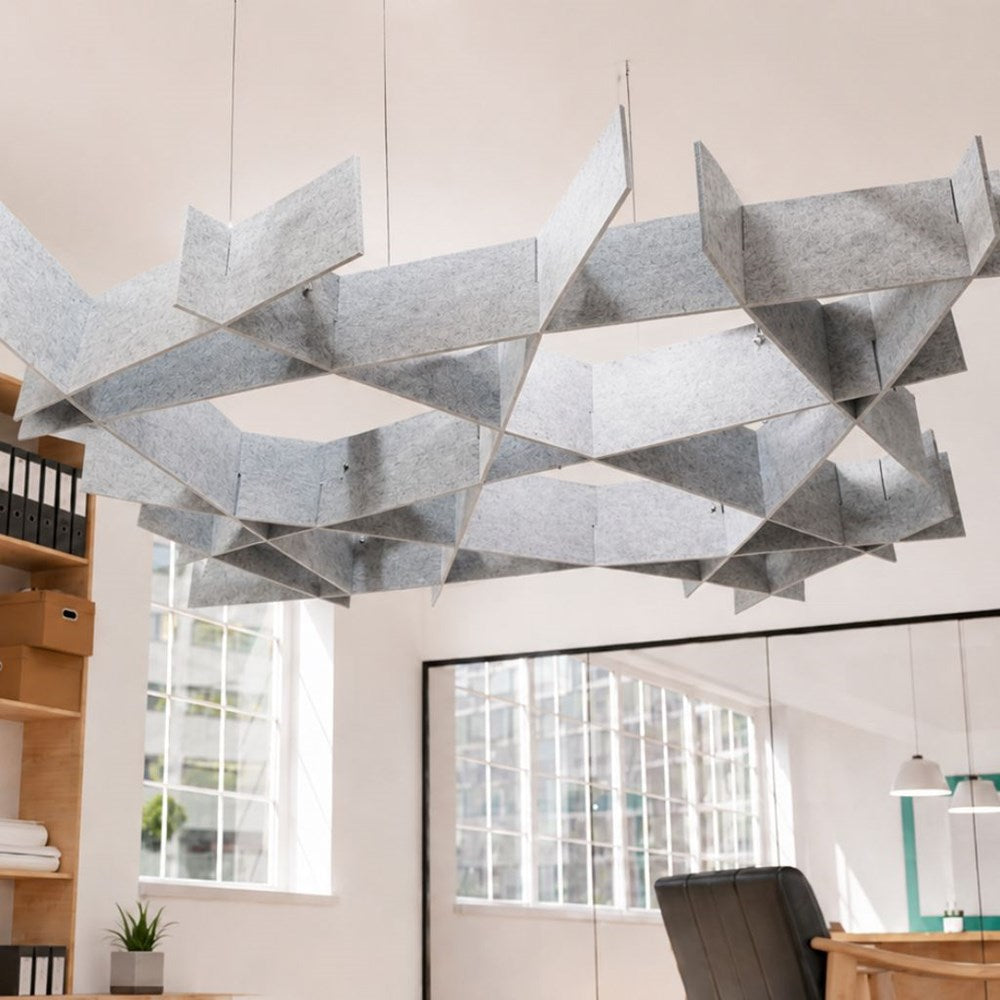 A large geometric ceiling light fixture made of layered silver-gray felt panels, suspended in a bright modern office space with windows, shelving, and a chair visible below.