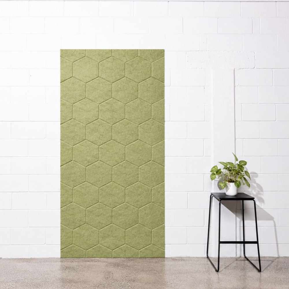 A tall rectangular panel of olive green hexagonal felt wall panels mounted on a white brick wall, with a small black side table and potted plant to the right in a minimalist room setting.