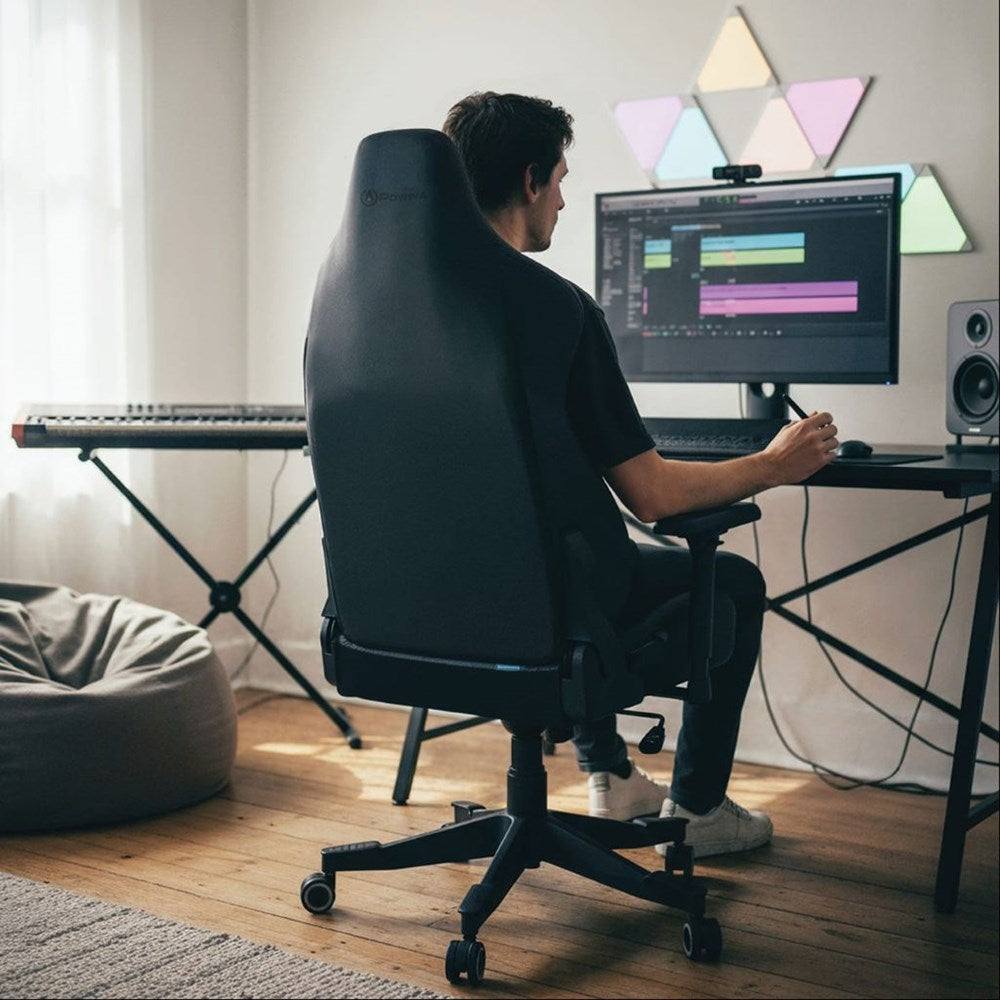 Person sitting at a desk with a computer, keyboard, and speakers in a room with geometric light fixtures.