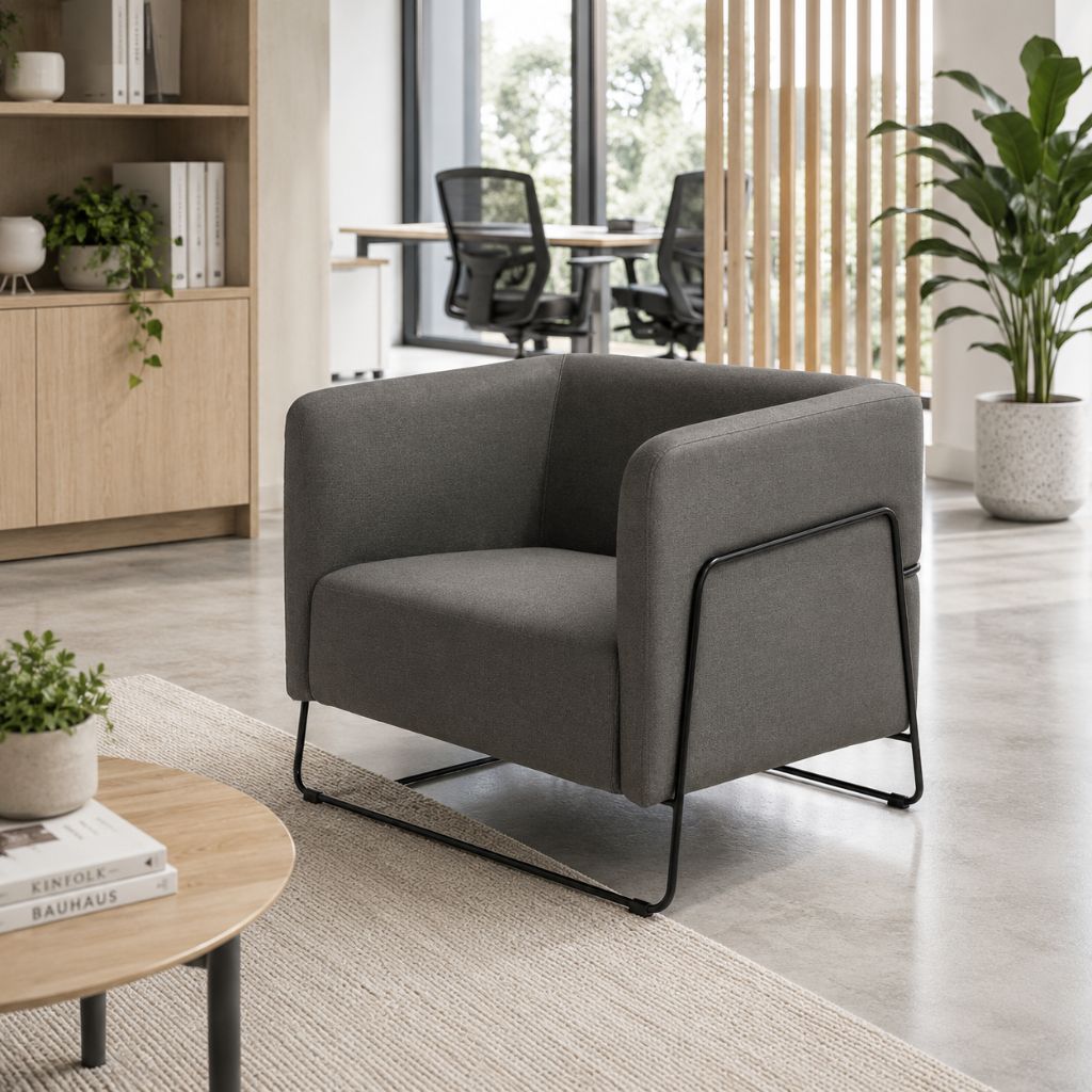 A modern gray upholstered accent chair with a black metal frame and legs, placed in a bright, contemporary office setting with a wooden table, shelving, and plants.