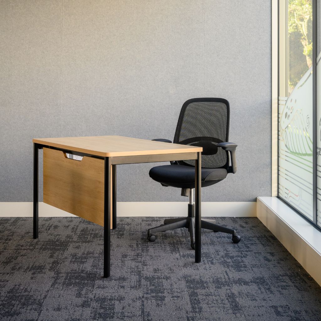 A modern teacher desk with a light wood top and front modesty panel, black metal legs, and a black mesh office chair, placed in a room with a gray textured wall, patterned gray carpet, and a large window on the right.
