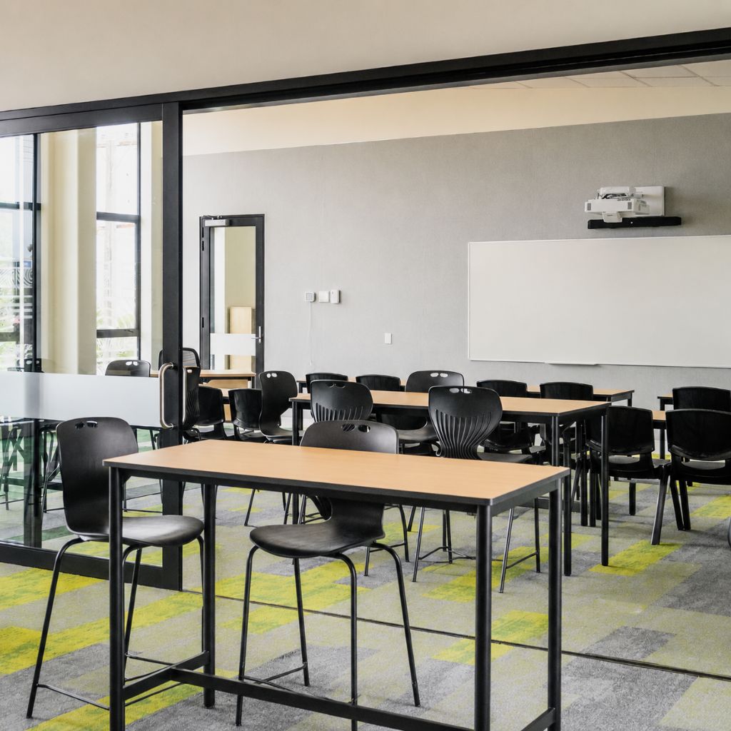 A modern classroom space with multiple black chairs, wooden-topped tables, a whiteboard, and a projector mounted on the wall. 
