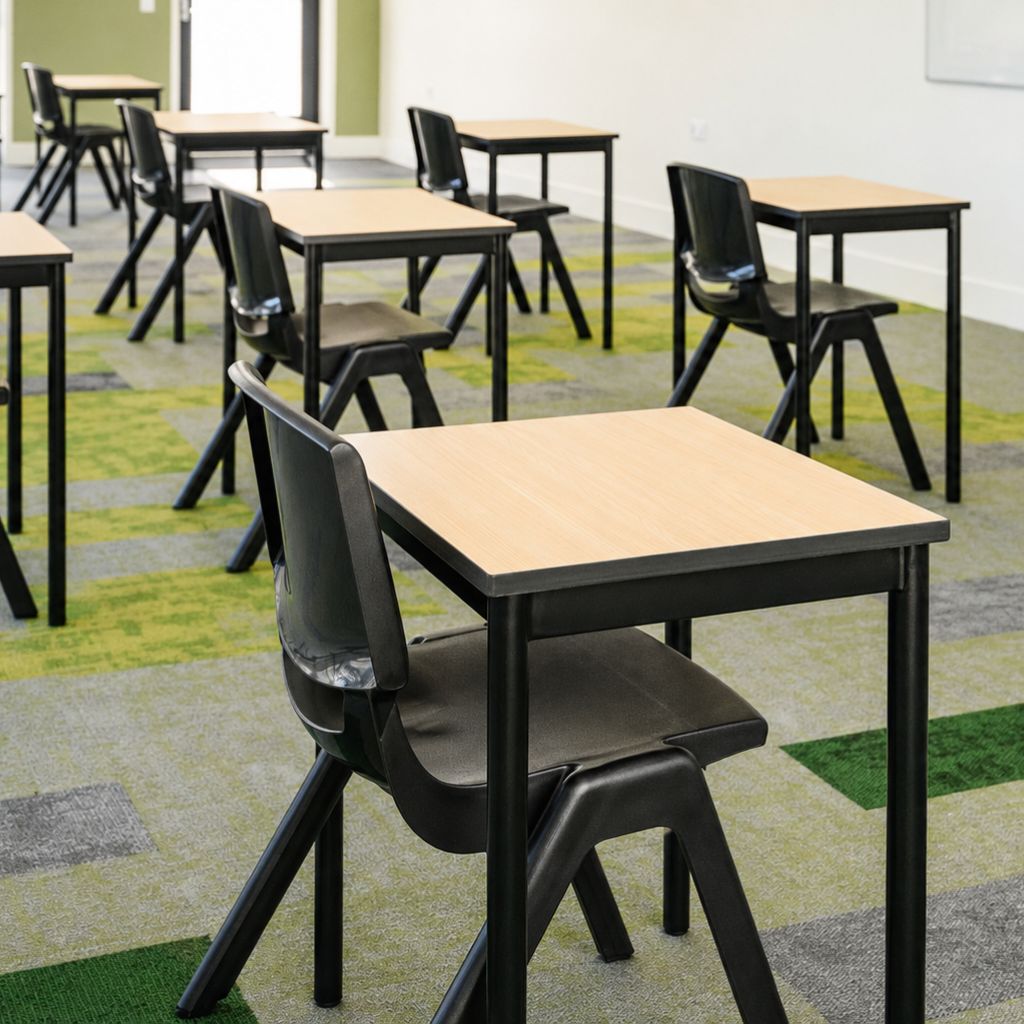 A modern classroom setup with black plastic chairs and matching black-framed tables with light wood tops, arranged on a green and gray patterned carpet.