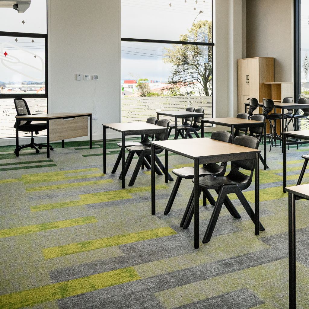 A modern classroom interior with multiple black chairs and light wood tables arranged on a gray and lime green patterned carpet. Large windows with black frames let in natural light, and a wooden cabinet is visible in the background.
